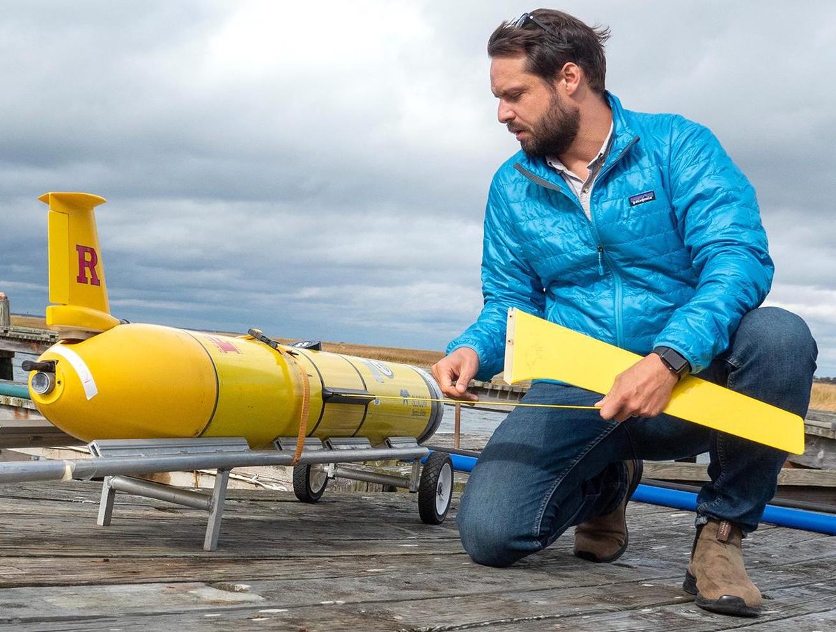Travis Miles kneeling on a dock beside a yellow Rutgers Slocum underwater glider